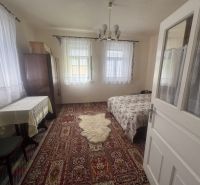 Ein Zimmer in einem Einfamilienhaus mit Teppich, Bett, Tisch und Vorhängen an den Fenstern.
