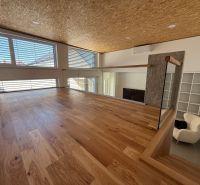 Innenraum eines Einfamilienhauses mit Holzboden und großen Fenstern.