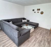 Wohnzimmer mit Ecksofa, Glastisch und Boden in Holzoptik.