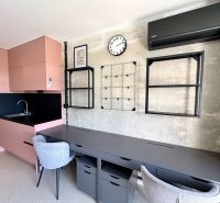 Ein Studio mit einem Boden in Holzoptik, einer rosa Küche und grauen Arbeitsstühlen.