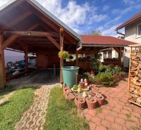 Ein Einfamilienhaus in Bacúch mit einem Unterstand, einem Garten und gelagertem Holz, an einem sonnigen Tag.