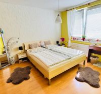 Schlafzimmer in einer Einzimmerwohnung mit Holzdekorboden, hellen Wänden, Fenstern mit weißen Vorhängen.