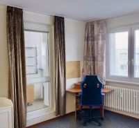 Einzimmerwohnung mit Fenstern, Vorhängen, Heizkörper und einem Tisch mit Stuhl.