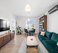Wohnzimmer in einer Zweizimmerwohnung mit einem großen Fernseher, einem Bücherregal und einem Boden in Holzoptik.