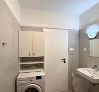 Badezimmer in einer 2-Zimmer-Wohnung mit Waschmaschine, weißem Schrank und Waschbecken auf hellen Fliesen.