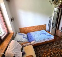 Ein Zimmer in einem Einfamilienhaus mit einem alten Teppich, einem Sessel und einem Bett am Fenster.