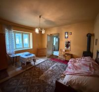 Innenraum eines Einfamilienhauses mit Bett, Tisch und Holzofen, in warmen Farbtönen gehalten.