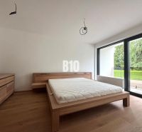 Schlafzimmer mit Bett und Kommode, Boden mit Holzdekor, großes Fenster mit Blick auf den Garten.