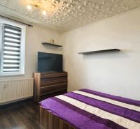 Schlafzimmer in einer 4-Zimmer-Wohnung mit Bett, Fernseher, Holzboden und modernen Jalousien.