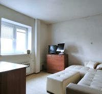 Ein Zimmer mit Sofa, Tisch, Holzelementen, Fernseher und Fenster mit Rollläden.
