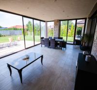 Ein verglaster Terrassenbereich eines Einfamilienhauses mit Blick auf den Garten und einem Boden aus Pflastersteinen.