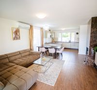 Wohnzimmer in einem Einfamilienhaus mit Küche, beigem Sofa und Boden in Holzoptik.