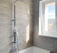 Badezimmer mit Badewanne und Dusche, Fliesen mit Holzdekor, großes Fenster, Einzimmerwohnung.