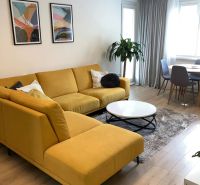 Wohnzimmer einer 3-Zimmer-Wohnung mit großem gelbem Sofa, Holzboden und Pflanze.