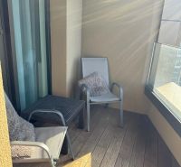 Balkon einer Zweizimmerwohnung mit Holzboden, zwei Stühlen und Glasgeländer.