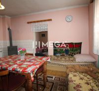 Innenraum eines Einfamilienhauses mit Küchentisch, Kachelofen und Bett, Boden mit Holzdekor.