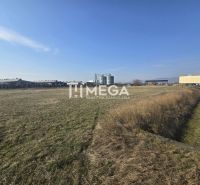 Gras und Produktionsräume in Hriadky unter klarem Himmel, mit einem Industriegebäude am Horizont.
