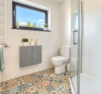 Badezimmer mit Dusche, Toilette und Fenster, bunte gemusterte Fliesen auf dem Boden.