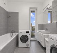 Badezimmer mit Badewanne, Waschmaschinen und Waschbecken in einer Zweizimmerwohnung. Graue Fliesen, großer Spiegel.