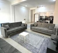 Wohnzimmer mit drei grauen Sofas, einem Couchtisch und einer Küche in einer 2-Zimmer-Wohnung.