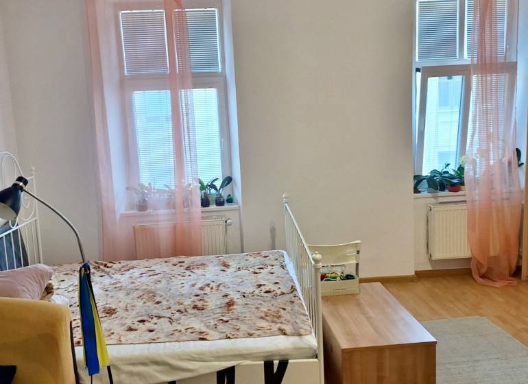 Innenraum einer Einzimmerwohnung mit Bett, Holzboden und Fenstern mit rosa Vorhängen.
