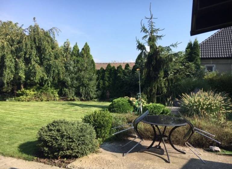 Der Garten des Einfamilienhauses in Čierna Voda mit Rasen, Bäumen und einer Außensitzgelegenheit.