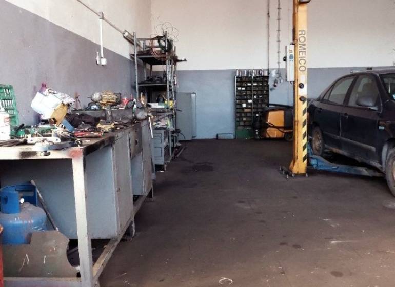 In der Werkstatt gibt es Werkzeuge, ein Auto auf dem Hebebühne und verstreute Teile auf dem Boden.