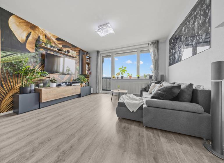 Wohnzimmer mit einem bequemen Sofa, einer Tapete mit Pflanzenmotiv und einem Boden in Holzoptik.