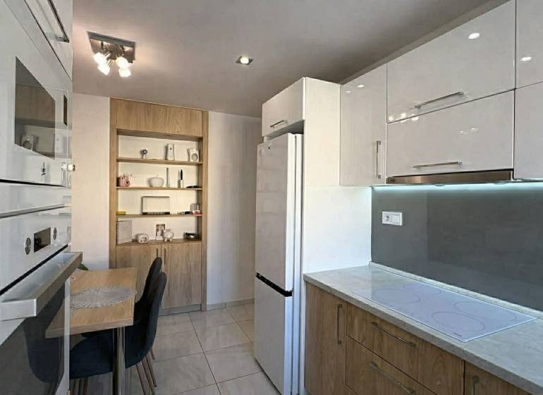 Küche in einer 2-Zimmer-Wohnung mit weißen Schränken, Tisch und Holzdekorregalen.