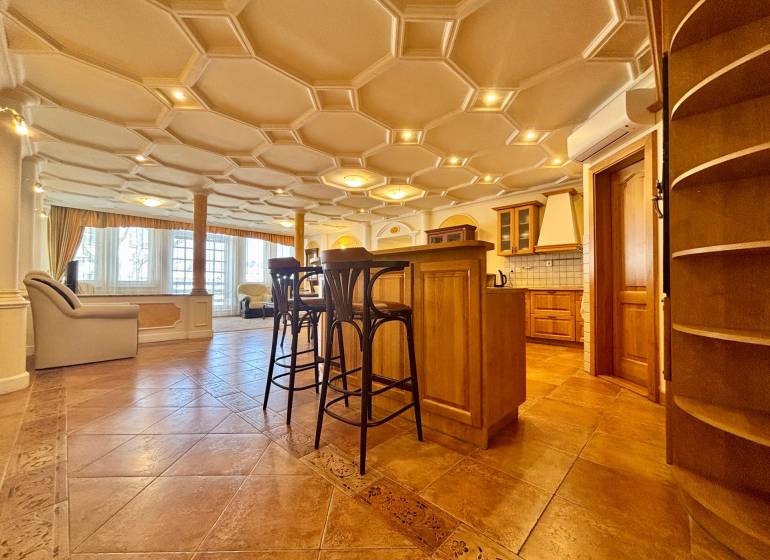 Innenraum einer 4-Zimmer-Wohnung mit dekorativer Decke, Holzboden, Kücheninsel und Stühlen.