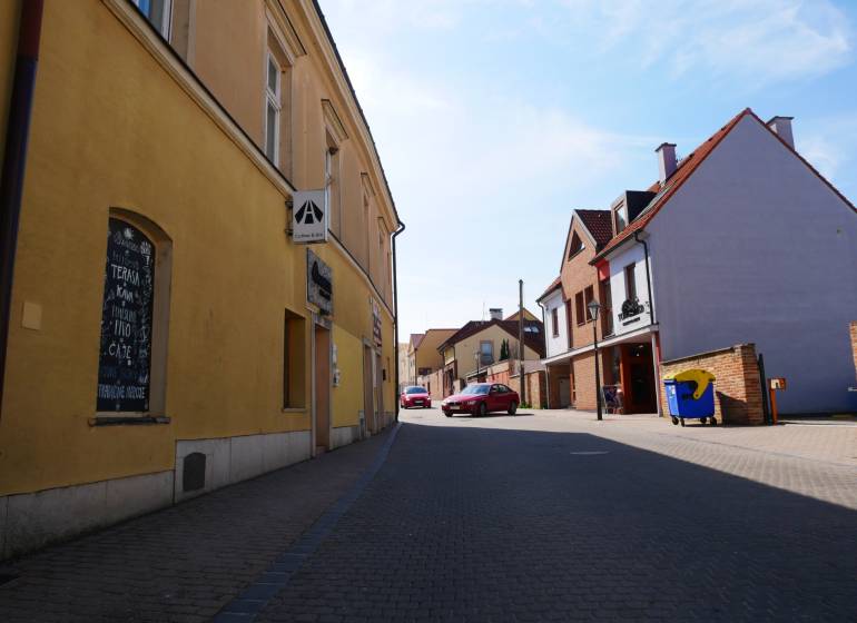 Eine Straße in Trnava mit Gastronomiebetrieben, gelbe und Backsteingebäude, geparktes Auto.