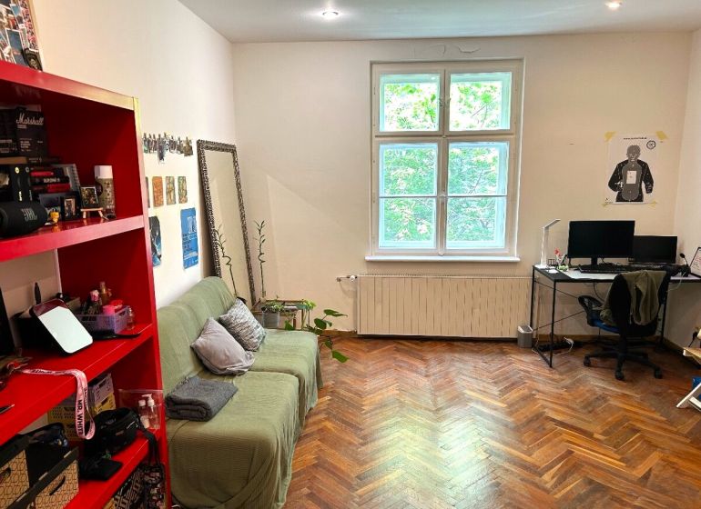Ein Zimmer in einer 2-Zimmer-Wohnung mit einem Boden in Holzoptik, einem Sofa, einem Schreibtisch und einem roten Regal.