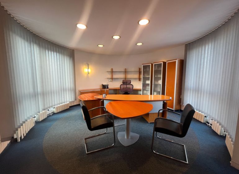 Ein Büro mit einem Tisch in Holzoptik, Regalen und beleuchteten vertikalen Jalousien.
