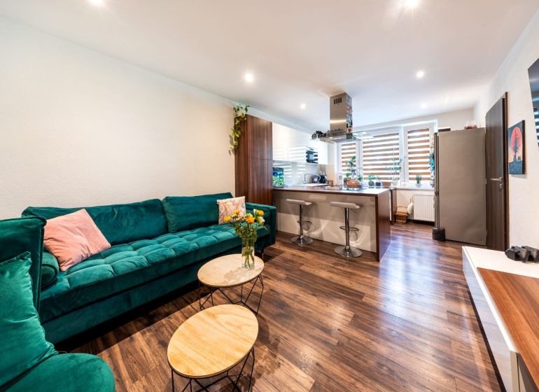 Wohnzimmer mit Küchenzeile in einer 3-Zimmer-Wohnung, Boden mit Holzdekor, grünes Sofa.