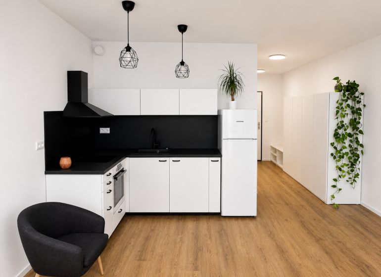 Küche in einer Einzimmerwohnung mit weißen Schränken, schwarzen Details und einem Boden in Holzoptik.