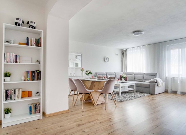 Wohnzimmer in einer Zweizimmerwohnung mit Holzboden, helle Sitzmöbel und Bücherregal.