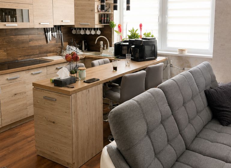 Wohnzimmer mit Küche, Holzboden, helle Schränke und graues Sofa.