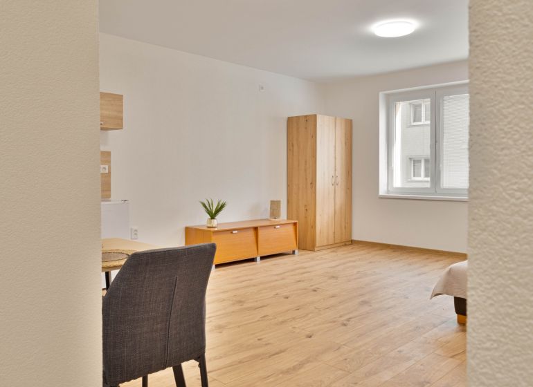 Einzimmerwohnung mit Holzboden, Schrank, Fenster, Tisch und zwei Schränkchen.