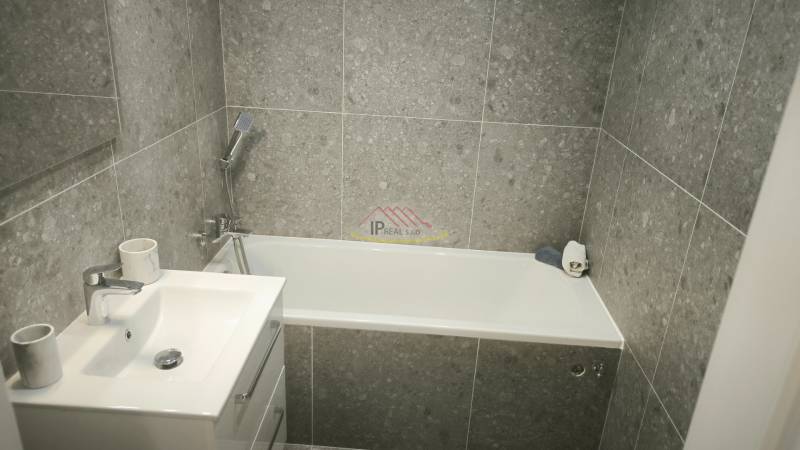 Badezimmer in einem Studio-Apartment mit Badewanne, Waschbecken und grauen Fliesen.