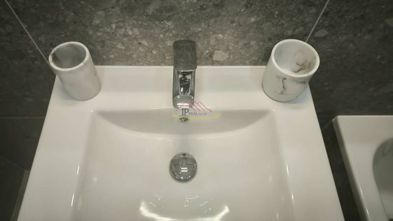 Weißes Waschbecken-Set in einem Studio-Apartment mit grauen Fliesen und einem Wasserhahn aus Metall.