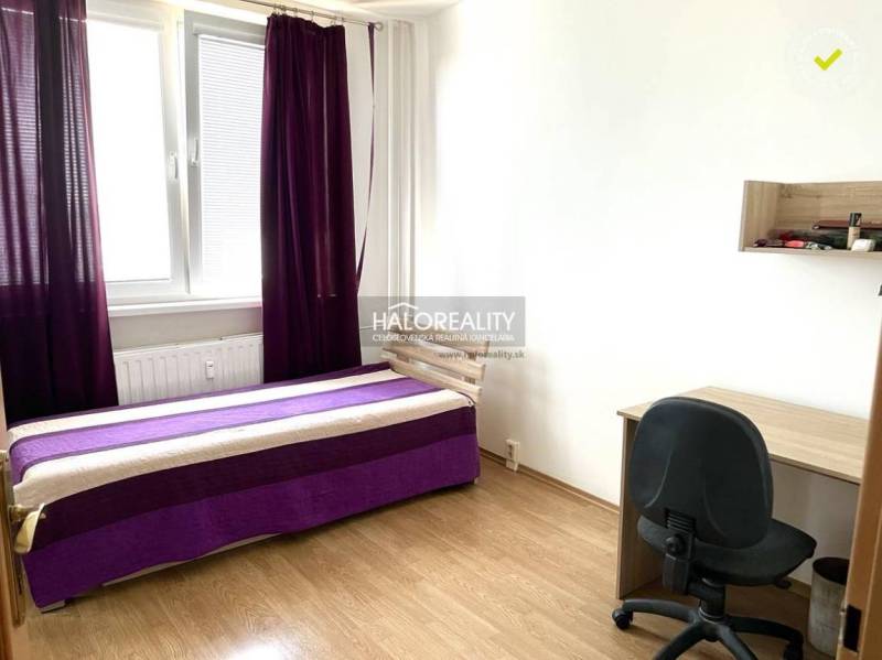 Zimmer: Bett mit lila Decke, Schreibtisch, schwarzer Stuhl, Fenster mit lila Vorhängen.