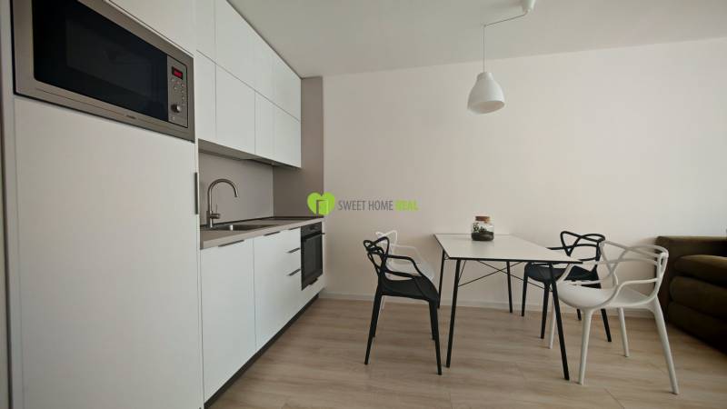 Küchenecke in einer Einzimmerwohnung, weiße Schränke, Tisch mit Stühlen, Holzboden.