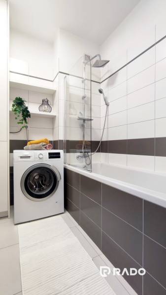Badezimmer in einer 2-Zimmer-Wohnung mit Waschmaschine, Badewanne, Regalen und einer dekorativen Pflanze.