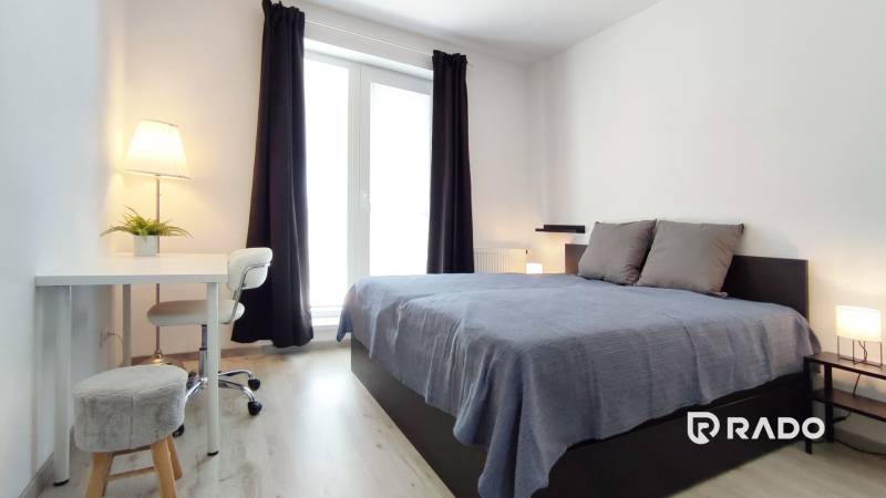 Helles Schlafzimmer in einer 2-Zimmer-Wohnung mit Bett, Tisch und Fenster mit Vorhängen.