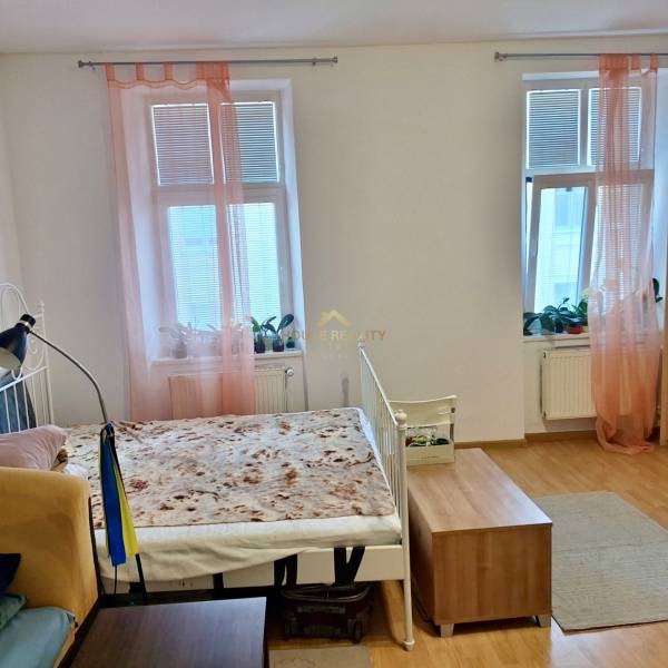 Innenraum einer Einzimmerwohnung mit Bett, Holzboden und Fenstern mit rosa Vorhängen.