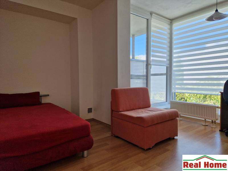 Ein Zimmer eines Einfamilienhauses mit einem roten Sofa und einem Sessel, großes Fenster mit horizontalen Jalousien.