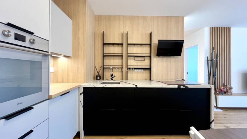 Küche in einer 2-Zimmer-Wohnung mit weißen und schwarzen Schränken, eingebautem Ofen und Holzelementen.