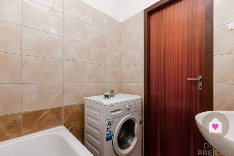 Badezimmer mit hellen Fliesen, Badewanne, Waschmaschine und brauner Tür in einer Einzimmerwohnung.