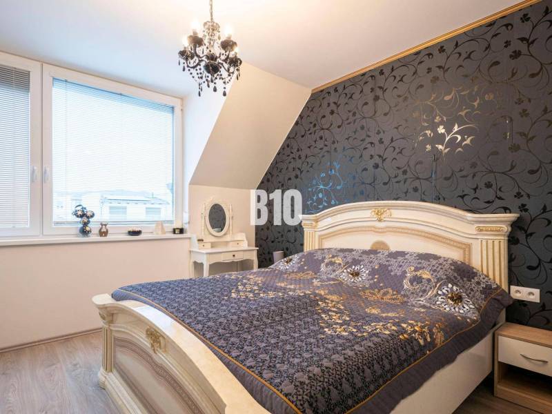 Ein Schlafzimmer in einem Einfamilienhaus mit dekorativer Tapete, elegantem Kronleuchter und Fenstern mit Jalousien.