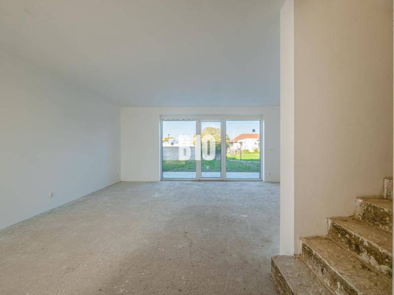 Ein Raum in einem Einfamilienhaus mit großen Fenstern und Betonboden, im Hintergrund eine Treppe.